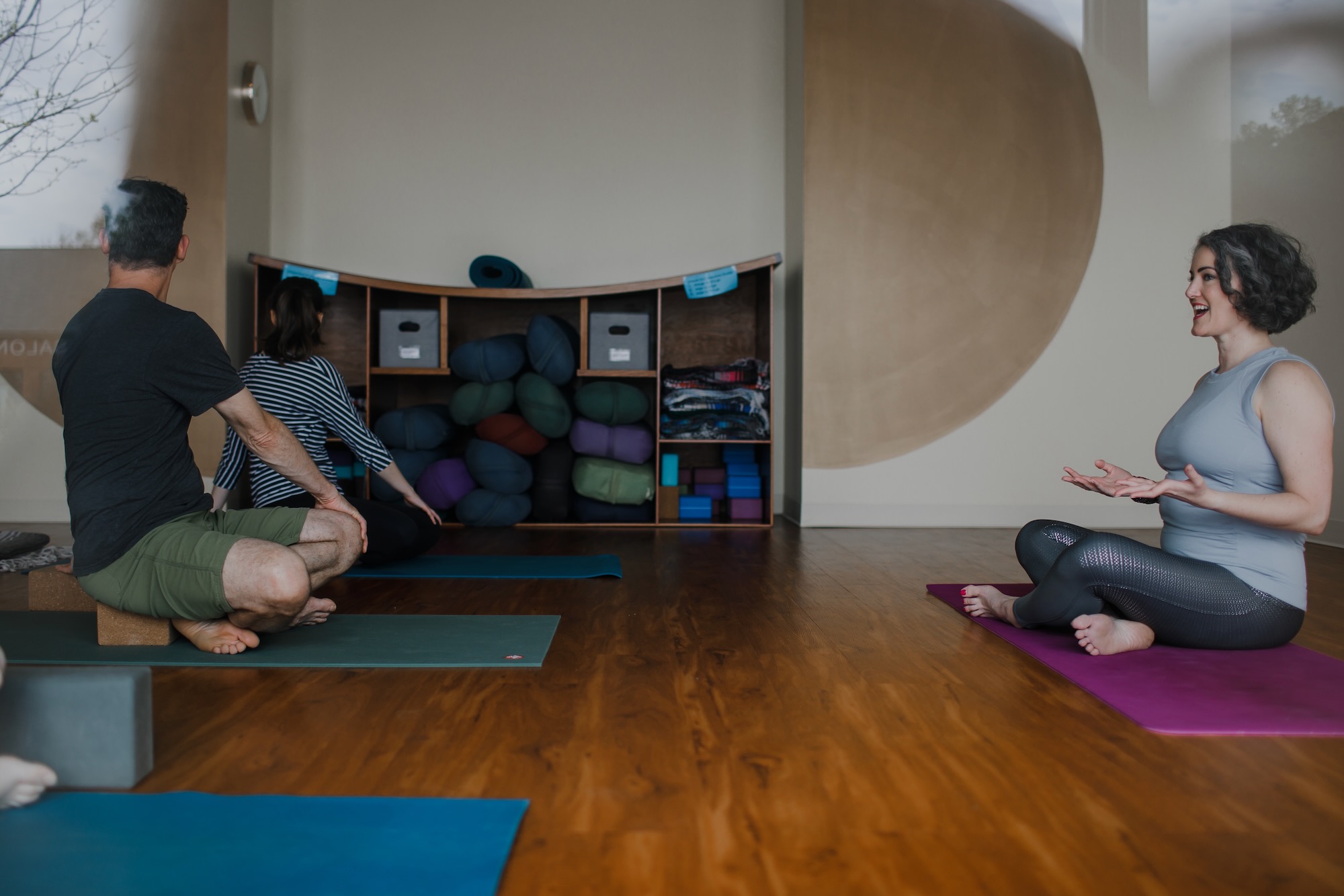 Sage sits at the front of the studio space talking as students do a seated twist